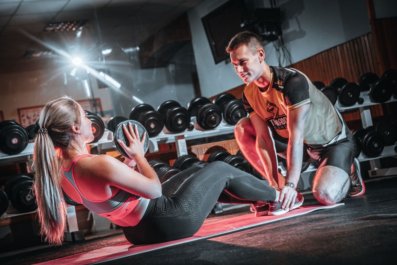 Home A personal trainer assists a woman doing sit-ups with a medicine ball in a gym setting.